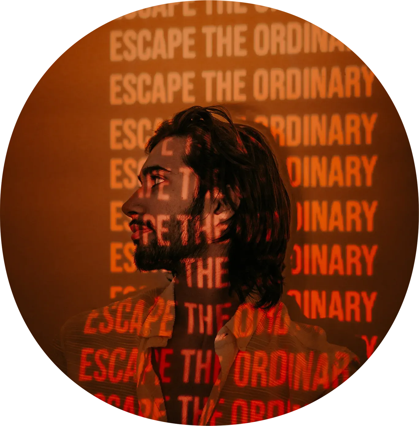 A man with long hair and a beard, looking to the side, with orange text that says 'ESCAPE THE ORDINARY' projected onto his face and background.
