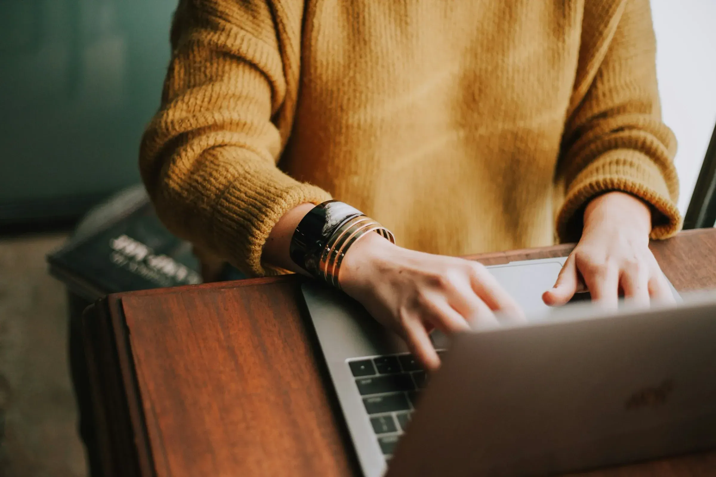 Person wearing a mustard-colored sweater using a laptop at a wooden desk.
