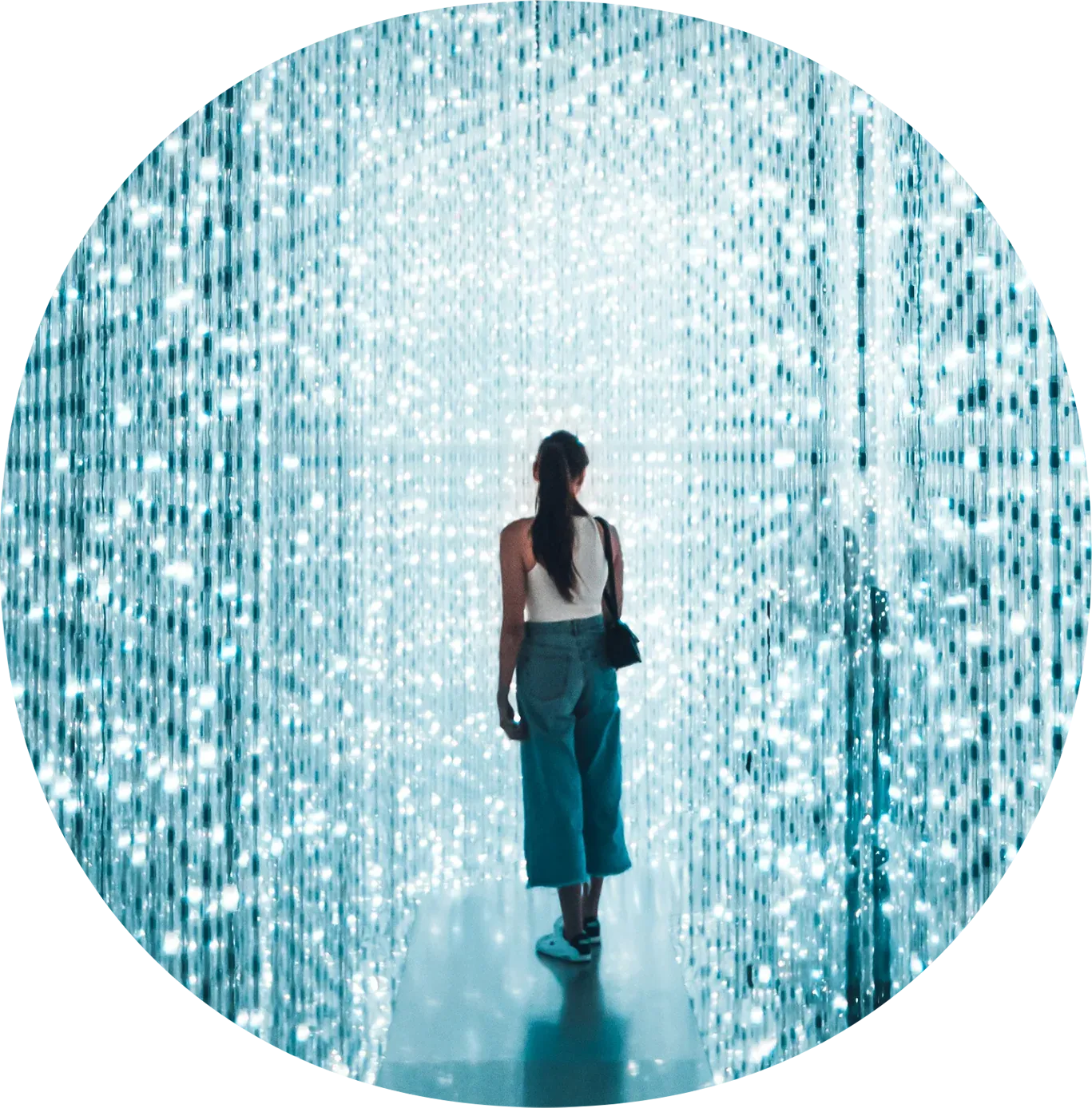 A woman with long dark hair in a white top and blue pants walking through an illuminated art installation with hanging LED lights.