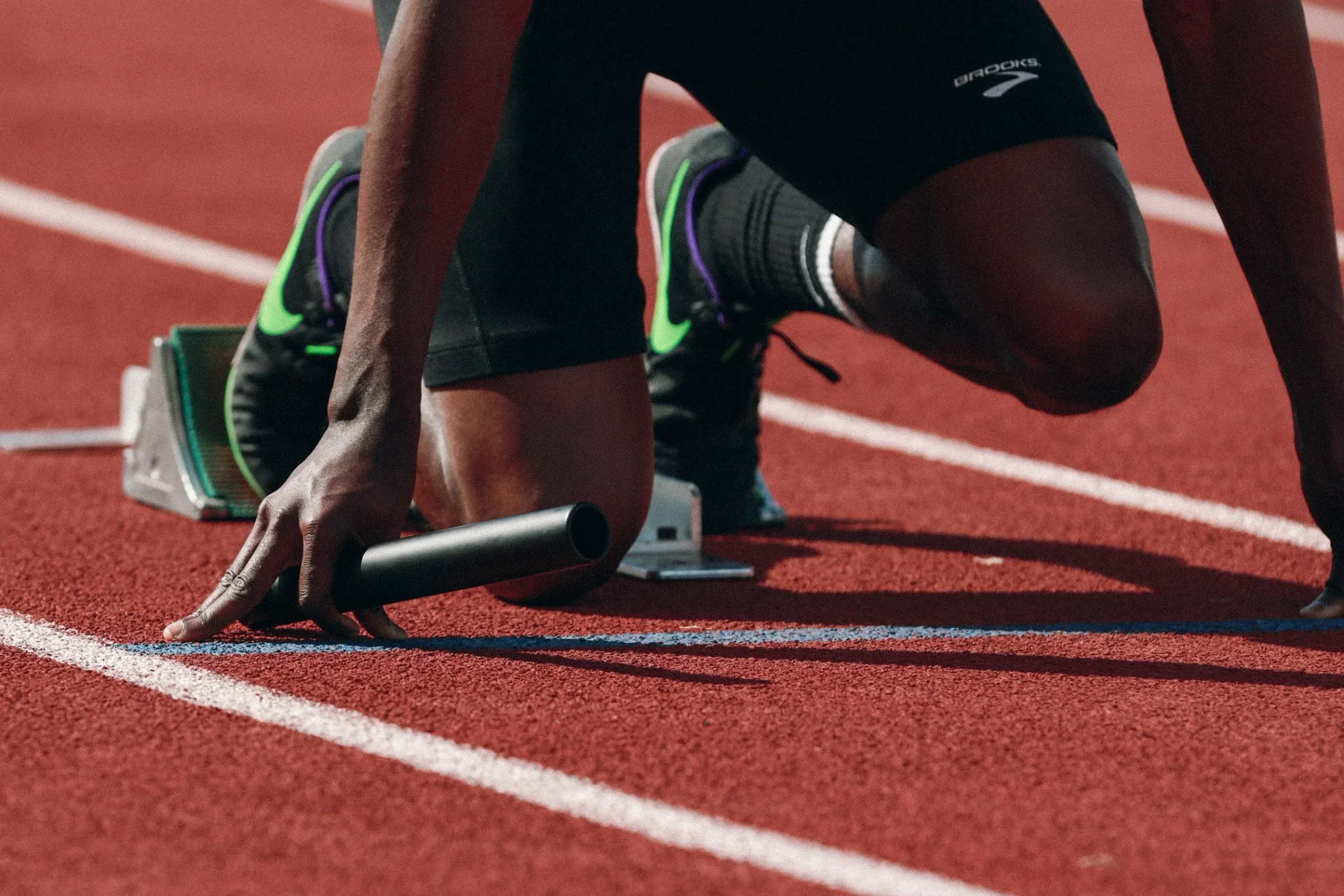 A sprinter in a starting position on a red track, wearing black athletic clothing and colorful running shoes, ready to race.