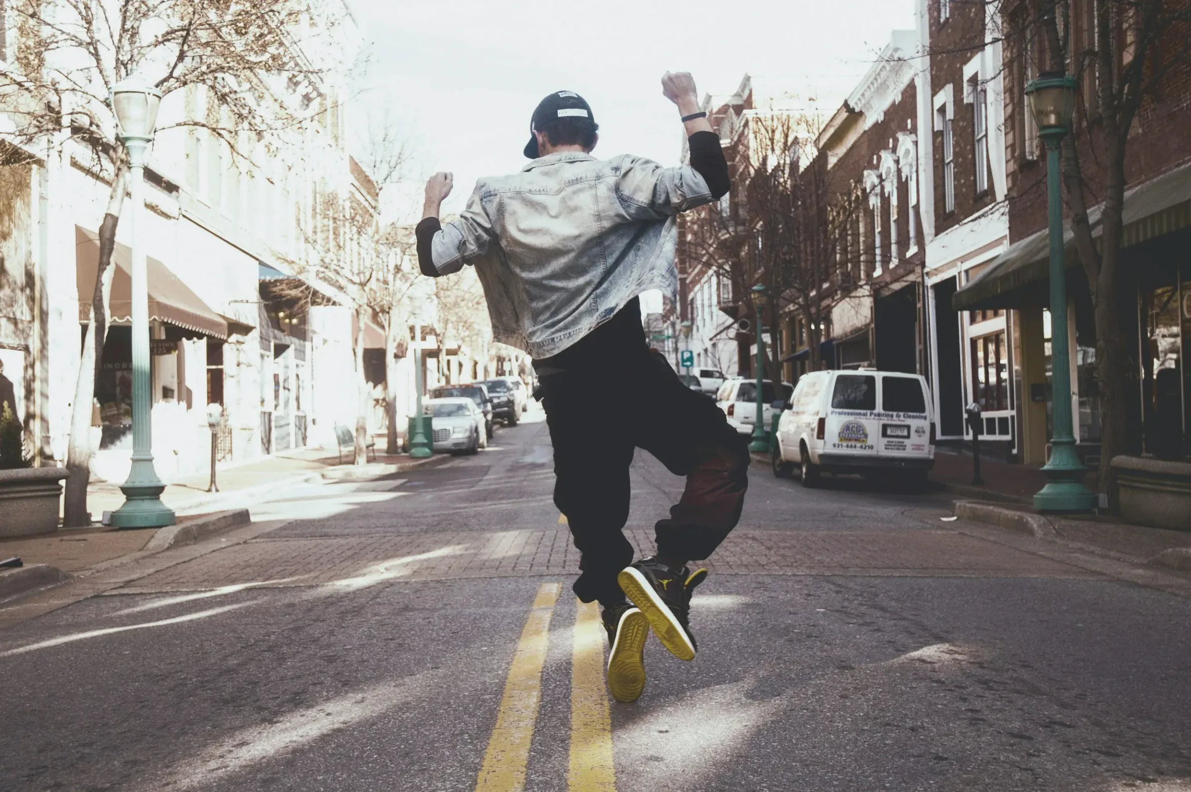 Young man dancing joyfully in the middle of a city street on a sunny day, with trees and parked cars visible in the background.