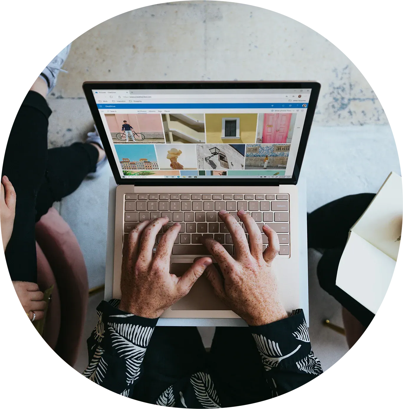 Person browsing an online photo gallery on a laptop, images of buildings and outdoor scenes visible on screen, person wearing black with patterned sleeves, sitting on light-colored floor with a white side table nearby.