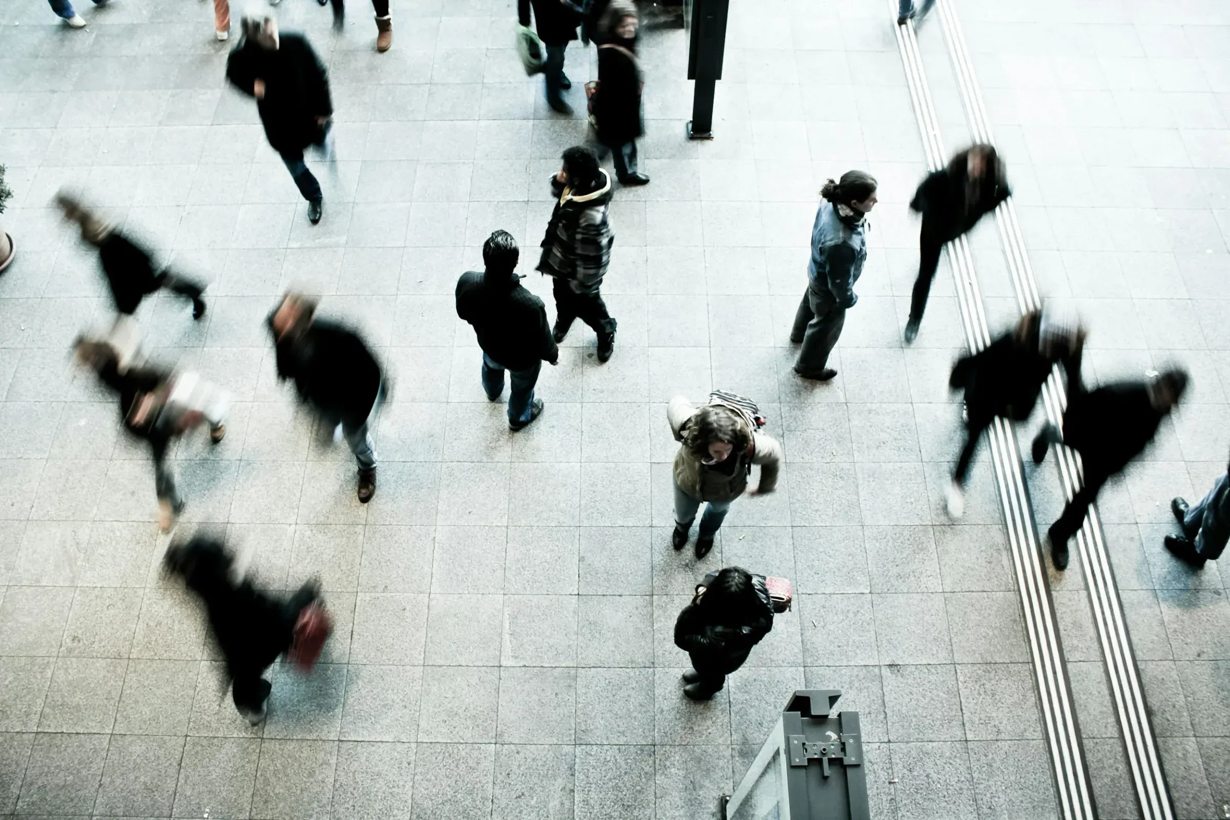 People walking on a tiled floor at a busy indoor venue, some blurred in motion, with stairs on the right side and a signpost in the lower part of the image.