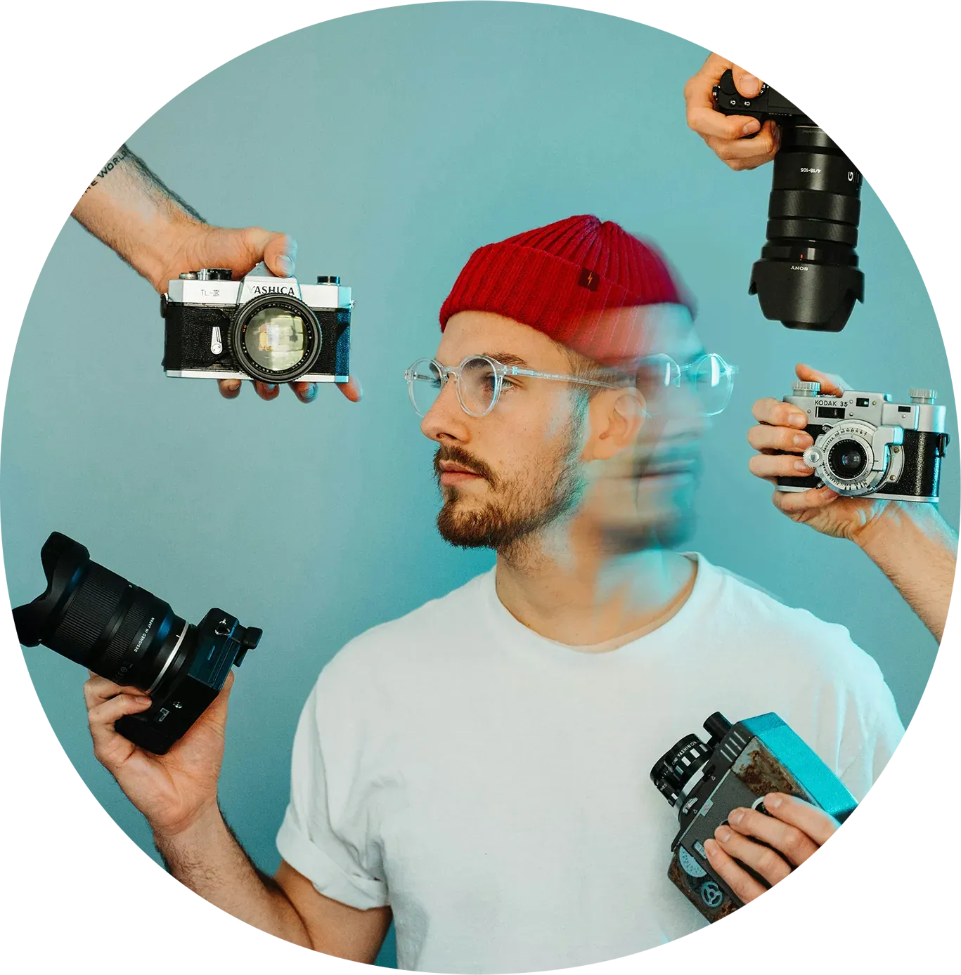 A man with glasses and a red beanie, holding a camera in each hand, while four other cameras are directed at him from different angles.