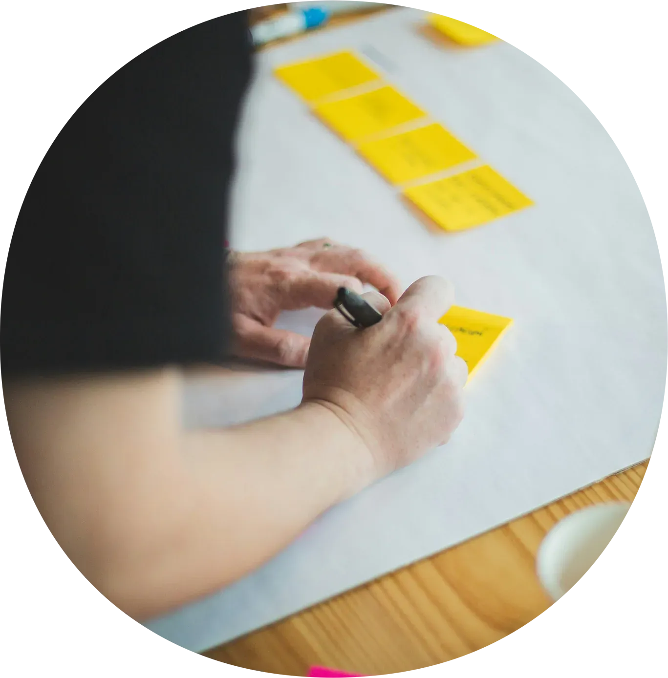 A person writing on a yellow sticky note at a table with additional sticky notes visible in the background.