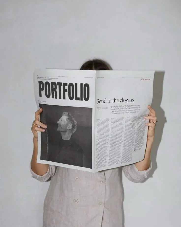 Person holding a newspaper with the headline 'PORTFOLIO' and a photo of a man laughing, standing against a plain white wall.
