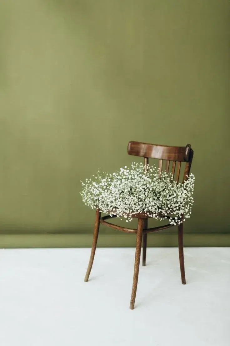 A wooden chair with a bouquet of small white flowers resting on its seat, set against a plain green background.